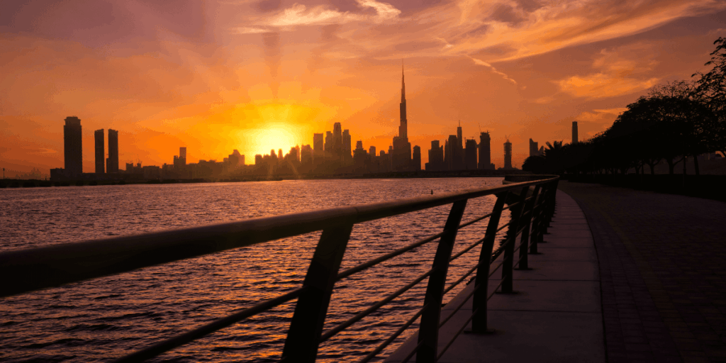 夕陽餘暉映照杜拜天際線，哈里發塔（Burj Khalifa）矗立於中央，橙紅色的天空與海面相互輝映，欄杆延伸的視角引導觀者進入畫面，營造出浪漫與沉靜的氛圍。