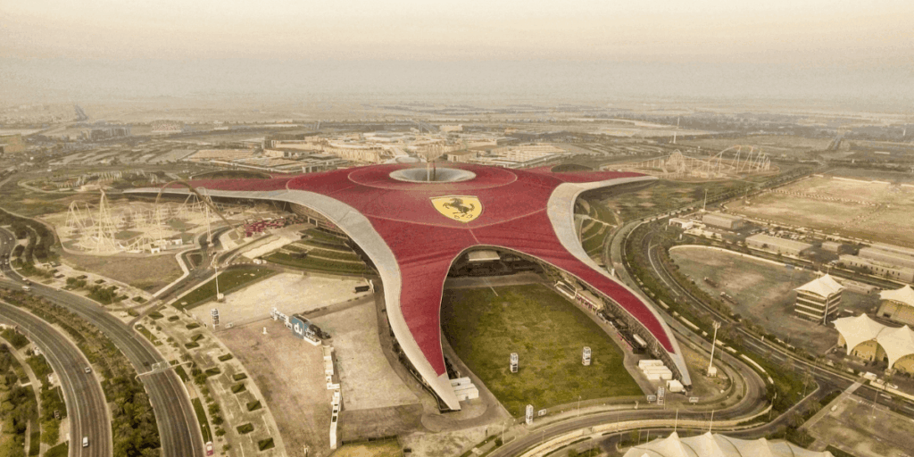 圖片以空拍角度呈現 Ferrari World 樂園，巨大紅色屋頂以法拉利標誌為中心向外延伸，外型猶如旋轉的星形，周圍可見雲霄飛車軌道與園區道路布局，是速度與冒險主題的象徵，也是深受家庭與遊客喜愛的熱門景點。