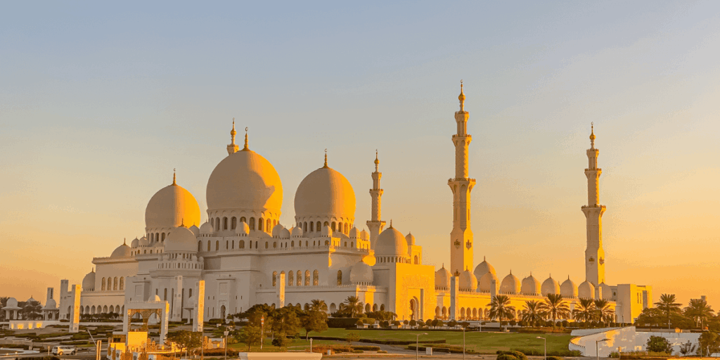 夕陽金光灑落在純白色的大清真寺上，圓頂與高塔展現細膩的伊斯蘭建築美學。建築在暖黃色光線下更顯神聖溫柔，是阿布達比最具代表性的宗教地標，也是旅人心目中的必訪拍照點。