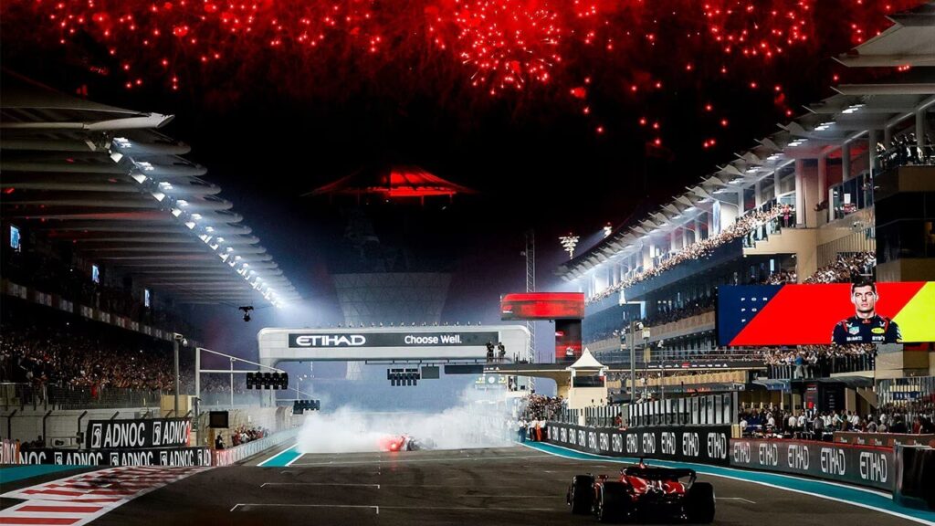 這張圖片呈現2024阿布達比亞斯碼頭賽道的F1夜間賽事畫面，夜空中綻放大量紅色煙火，照亮整個賽道上方。畫面中央可見終點線位置，賽道左、右兩側的看台擠滿觀眾，燈光與螢幕營造出濃厚賽事氛圍。前方有F1賽車高速駛過，伴隨煙霧與燈光，展現賽季最終站壓軸的激情與震撼，是極具視覺張力的比賽場景。